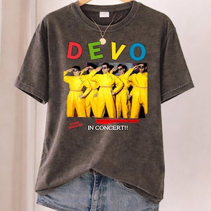 May include: Dark gray t-shirt featuring the band name "DEVO" in colorful letters above a graphic of the band members in yellow jumpsuits. The text "IN CONCERT!!" is below the graphic. A gold necklace hangs on the shirt.