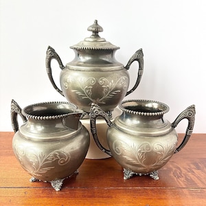 May include: A silver tea set with a teapot and two serving bowls. The set has ornate handles and floral engravings. The teapot has a lid with a decorative finial. The set is displayed on a wooden surface.