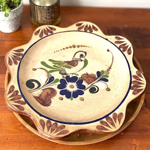 May include: A decorative ceramic plate with a scalloped edge, featuring a hand-painted bird and floral design in brown, blue, and green. The plate rests on a wooden charger, showcasing a rustic, artisanal aesthetic.