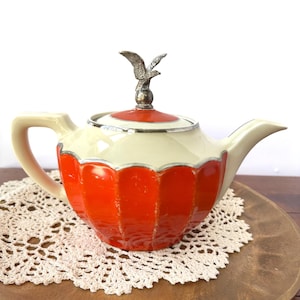 May include: A vintage teapot in cream and orange. The body is cream-coloured with an orange lower section. It has a silver trim and an eagle finial. The teapot has a curved handle and spout, and sits on a crocheted doily.