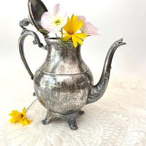 May include: A vintage silver-plated teapot with an open lid, filled with yellow and pink flowers. The teapot has a curved spout, ornate handle, and rests on three decorative feet. The background is a white lace tablecloth.