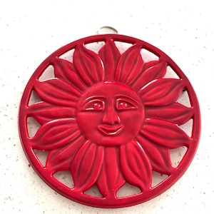 May include: A round, red trivet with a smiling sun face design. The trivet features a detailed sunflower pattern with a circular border. This decorative item is likely used to protect surfaces from hot cookware.