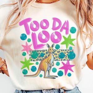 May include: Cream-colored t-shirt with a kangaroo graphic. The design includes the words "Too Da Loo" in pink, along with teal polka dots, green and pink stars, and a leopard print stripe. The kangaroo is tan and yellow.