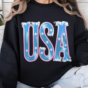 Può includere: Felpa nera con la scritta "USA" in grandi lettere stilizzate. Le lettere sono blu con un contorno rosa e dettagli di stelle bianche, creando un design patriottico. Un capo casual.