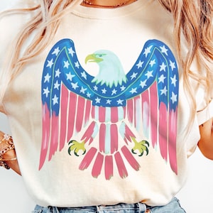 May include: Cream-coloured t-shirt with a patriotic eagle design. The eagle has blue wings with white stars, red and white striped body, and a light blue head with a yellow beak and talons. The design is painted.