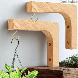 May include: Two wooden plant shelf brackets with brass hooks. The brackets are made of light-coloured wood and have a curved design. One bracket has a hanging plant with green and yellow leaves. The brand name "Nook & Alder" is in the top right corner.