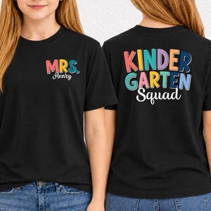 Può includere: Una t-shirt nera con una grafica multicolore sul petto sinistro che recita MRS. Henry. Il retro della maglietta mostra il testo multicolore KINDERGARTEN Squad in un carattere audace e giocoso. Il design utilizza rosa, giallo, blu e arancione.