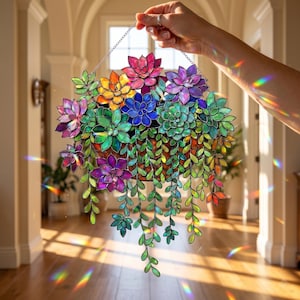 May include: A stained glass suncatcher featuring a collection of multi-colored succulents in a hanging basket. The piece displays vibrant shades of pink, purple, orange, blue, and green glass segments with dark metal soldering, creating light refractions.
