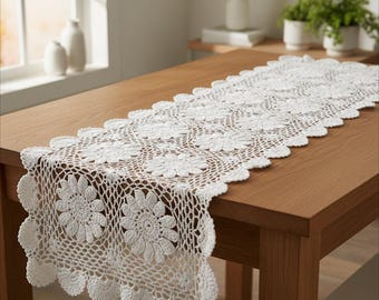 May include: A white, crocheted table runner with a floral pattern. The runner is placed on a wooden table. The runner's dimensions are approximately 60 inches long and 14 inches wide.