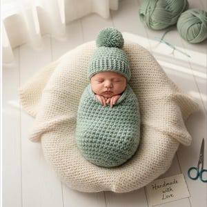 Può includere: Un neonato avvolto in un bozzolo e cappello all'uncinetto verde menta con un pompon. Il bambino riposa su una coperta di maglia color crema. La scena include filato e forbici, suggerendo un articolo fatto a mano.