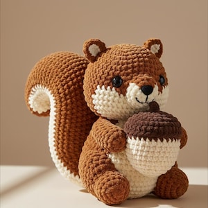 Puede incluir: Un peluche de crochet con forma de ardilla, en tonos marrón, crema y blanco, sosteniendo una bellota marrón y crema. La ardilla tiene ojos negros y una cola esponjosa. Un juguete suave y hecho a mano.