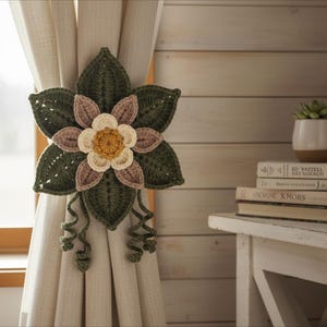 May include: A handmade, crochet curtain tieback in the shape of a flower. The flower has dark green leaves, with petals in shades of beige, light brown, and yellow. The tieback is attached to a beige curtain.