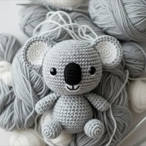 May include: A handmade grey crochet koala bear with a black nose and small black eyes. The koala is surrounded by grey yarn, with white yarn accents. The koala has white inner ears and a smiling mouth.