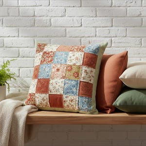 Peut inclure: Un coussin carré en patchwork avec un motif floral dans les tons de rouge, bleu et vert. Il est posé sur une surface en bois avec d'autres coussins unis. Une petite plante et une couverture pliée sont également visibles.