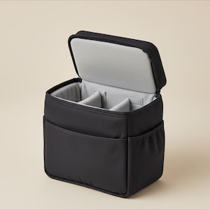 May include: Black camera bag with an open lid, revealing a gray interior with dividers. The bag has exterior pockets on the front and sides. The bag is designed to protect and organize camera equipment.