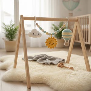 Könnte beinhalten: Ein Holz-Spielbogen für Babys mit hängenden Häkeltieren, darunter eine lächelnde Wolke, eine Sonne und ein Heißluftballon. Die Spielzeuge sind in sanften Pastellfarben gehalten. Eine graue Decke und eine Holzrassel liegen auf einem flauschigen weißen Teppich.