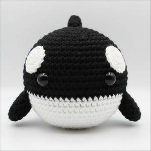 May include: A handmade crochet orca whale plushie. The plush toy features a black body with white markings, including eye spots and a white belly. It has small black button eyes and a dorsal fin. The orca is a cute, soft toy.