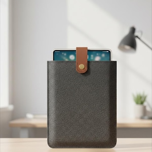 May include: A dark gray tablet sleeve with a brown leather strap and gold button closure. The tablet is partially visible inside the sleeve. The sleeve is made of a textured material and is designed to protect the tablet.