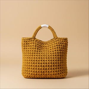 Può includere: Borsa tote all'uncinetto giallo senape con la parte superiore arrotondata e due manici. I manici sono collegati da una fascia bianca. La borsa è realizzata con filato spesso e ha un aspetto strutturato. La borsa è su uno sfondo neutro.
