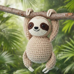 Puede incluir: Un juguete de perezoso tejido a ganchillo en beige, crema y marrón cuelga de una rama marrón sobre un fondo verde borroso. El perezoso tiene una cara color crema con parches marrones en los ojos y una sonrisa negra cosida. Sus brazos y piernas están extendidos, con garras blancas.