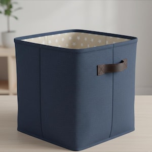 Puede incluir: Un cubo de almacenamiento azul marino con forma cuadrada y un asa de cuero marrón. El interior está forrado con tela beige con patrones de estrellas blancas. Diseñado para organizar objetos.