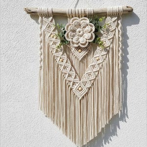 May include: Cream-coloured macrame wall hanging featuring a central flower design and wooden beads. It is suspended from a natural driftwood branch. Greenery accents the flower, adding a touch of nature to this handcrafted decor.