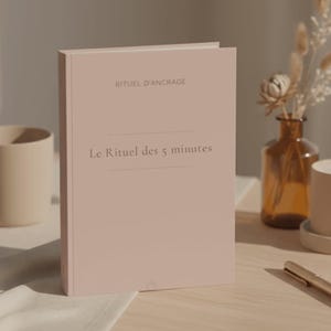 May include: A soft pink hardcover book, titled "RITUEL D'ANCRAGE" and "Le Rituel des 5 minutes" in a minimalist font. The book is on a light wooden surface, with a blurred background featuring a vase and a pen.