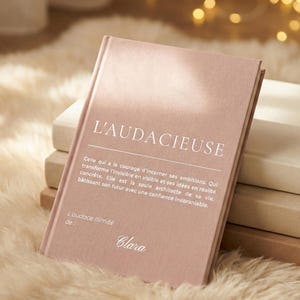 Puede incluir: Un diario rosa con el texto "L'AUDACIEUSE" en blanco. El diario tiene una cita en francés y el nombre "Clara" en la parte inferior. Está sobre una superficie suave y clara.