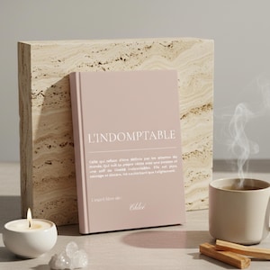 Puede incluir: Un libro rosa titulado "L'INDOMPTABLE" con texto en francés, junto a una vela encendida, una taza de té con vapor y un pequeño cristal. El libro se apoya en un bloque de piedra texturizada. Palos de santo están sobre la mesa.