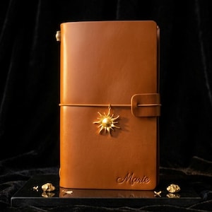 Peut inclure: Une couverture de journal en cuir beige avec une breloque dorée en forme de soleil attachée à une sangle de fermeture élastique horizontale. Le nom Marie est embossé dans le coin inférieur droit. La couverture a une texture lisse et une boucle.