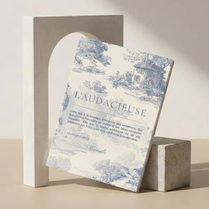 Puede incluir: Un libro de tapa dura titulado "L'AUDACIEUSE" con un diseño de toile de Jouy azul y blanco. El libro está apoyado en un pequeño cubo gris, con una estructura blanca en forma de arco en el fondo. La portada del libro presenta un paisaje.