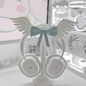 May include: White over-ear headphones resting on a white stand. The headband is decorated with a pair of white angel wings and a light blue bow centered between them. The headphones feature a small silver logo on each of the two ear cups.
