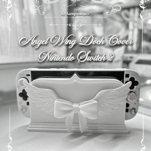 May include: A white Nintendo Switch dock cover featuring a central bow and sculpted angel wings on the front. The text 'Angel Wing Dock Cover Nintendo Switch 2' and 'by kissesfrmangels' is displayed above the device in a decorative font.