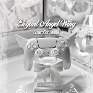 May include: A white controller stand featuring sculpted angel wings on the sides and a prominent bow on the front. The stand holds a white game controller. Text on the image reads Elegant Angel Wing controller stand and by kissesfromangels.