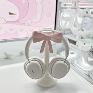 Peut inclure: Casque audio sans fil blanc avec un support assorti, surmonté d'un nœud rose clair. Le casque repose sur un support blanc, créant une esthétique épurée. L'arrière-plan présente un écran d'ordinateur avec une image de dessin animé rose.