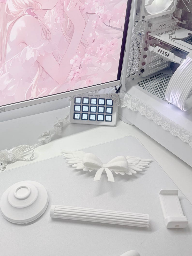 May include: A white desk setup featuring a central 3D-printed white bow with feathered wings, a circular white base, a cylindrical fluted rod, and a rectangular phone stand. In the background, a computer monitor displays pink anime art.