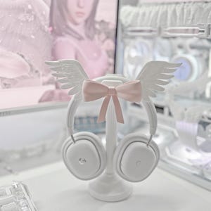 May include: A white headphone stand featuring a pair of white angel wings attached to the top arch and a light pink bow centered between the wings. The stand holds a pair of white over-ear headphones with a subtle logo on each ear cup.
