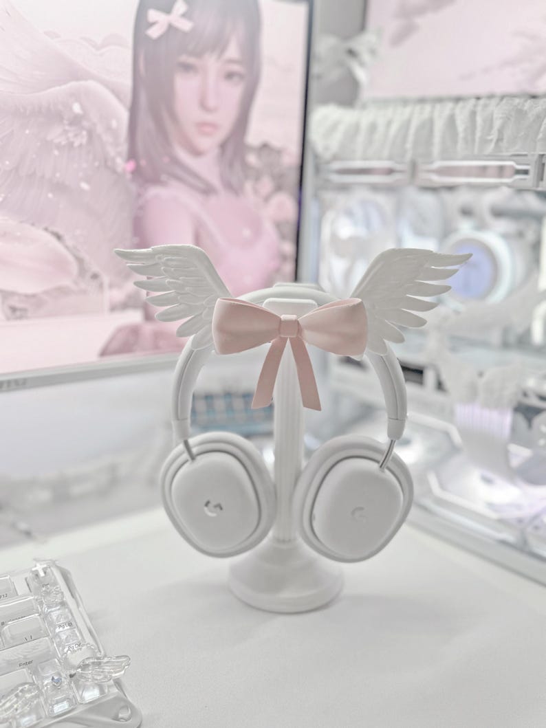 May include: A white headphone stand featuring a pair of white angel wings attached to the top arch and a light pink bow centered between the wings. The stand holds a pair of white over-ear headphones with a subtle logo on each ear cup.