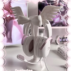 May include: A white headphone stand featuring a central pillar base, adorned with a large bow and a pair of sculpted angel wings at the top. The stand holds a pair of white and rose gold over-ear headphones with a microphone attached to the left side.
