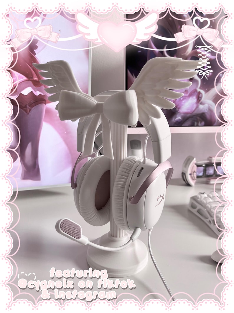 May include: A white headphone stand featuring a central pillar base, adorned with a large bow and a pair of sculpted angel wings at the top. The stand holds a pair of white and rose gold over-ear headphones with a microphone attached to the left side.