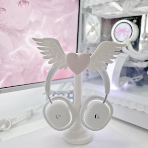 Puede incluir: Unos auriculares inalámbricos blancos sobre un soporte, con un adorno en la diadema que consiste en un corazón rosa claro central flanqueado por dos alas blancas con plumas. Los auriculares tienen un acabado liso y un pequeño logotipo.