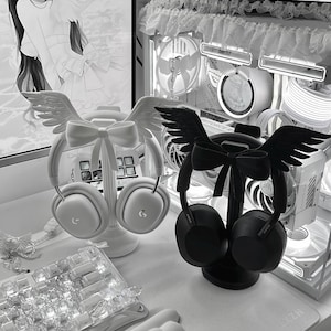 May include: Two headphone stands, one white and one black, each featuring a decorative bow and a pair of angel wings at the top. The white stand holds white over-ear headphones, while the black stand holds black over-ear headphones.