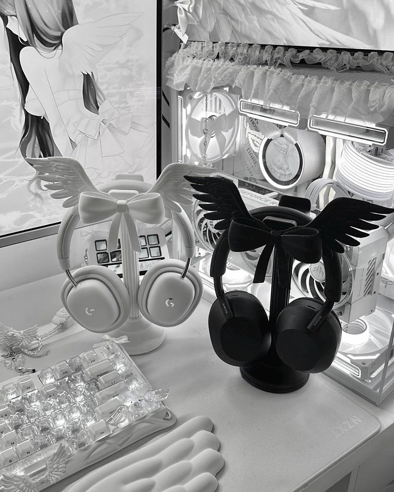 May include: Two headphone stands, one white and one black, each featuring a decorative bow and a pair of angel wings at the top. The white stand holds white over-ear headphones, while the black stand holds black over-ear headphones.