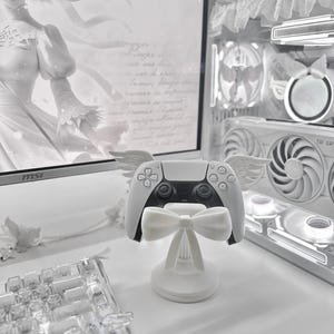 Pode incluir: Um comando branco PlayStation 5 numa base em forma de laço com detalhes de asas. O comando tem botões pretos. A base é branca e apresenta um design de laço. O fundo inclui um monitor de computador, um teclado e componentes de computador.