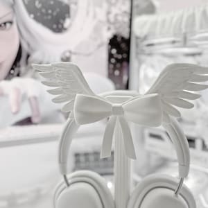 May include: A pair of white over-ear headphones featuring a decorative attachment on the headband. The attachment consists of a central bow flanked by two symmetrical feathered wings. The headphones have square-shaped ear cups with a small logo.