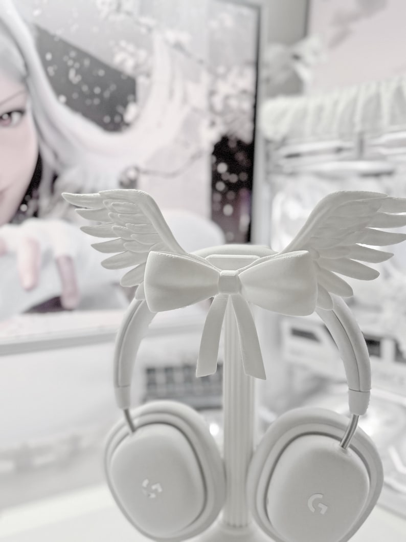 May include: A pair of white over-ear headphones featuring a decorative attachment on the headband. The attachment consists of a central bow flanked by two symmetrical feathered wings. The headphones have square-shaped ear cups with a small logo.