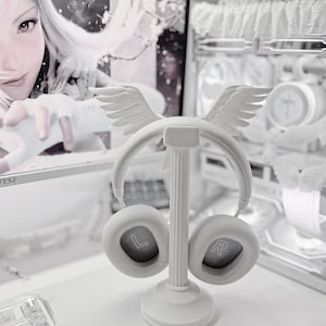 May include: A white headphone stand featuring a central column with a circular base and a top ornament shaped like a pair of wings. The stand holds a pair of white over-ear headphones with the letters L and R clearly visible inside the ear cups.