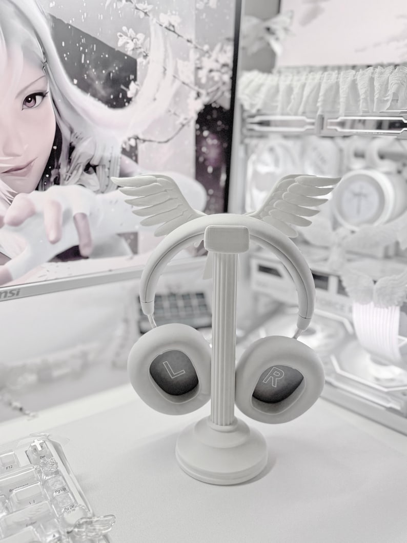 May include: A white headphone stand featuring a central column with a circular base and a top ornament shaped like a pair of wings. The stand holds a pair of white over-ear headphones with the letters L and R clearly visible inside the ear cups.