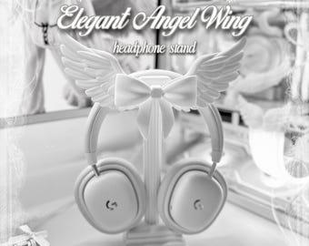 Puede incluir: Un soporte para auriculares blanco con un pilar central vertical, una base y un elemento decorativo superior en forma de lazo flanqueado por dos alas de ángel emplumadas. El texto "Elegant Angel Wing headphone stand" y "by kissesfrmangels" es visible.