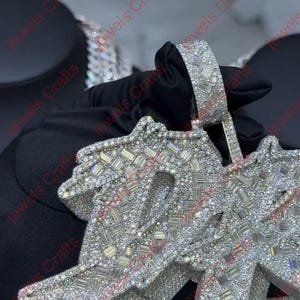 May include: A large, silver-toned pendant with a stylized design, heavily encrusted with sparkling, clear gemstones. The pendant is held by a gloved hand, showcasing the intricate details and craftsmanship of the jewelry.